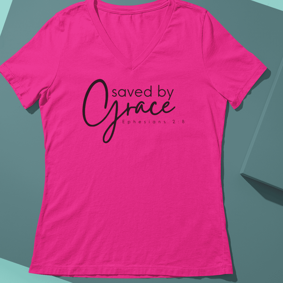 Image shows a pink v neck t shirt that says Saved By Grace Ephesians 2:8. Design by Gathered-Sown